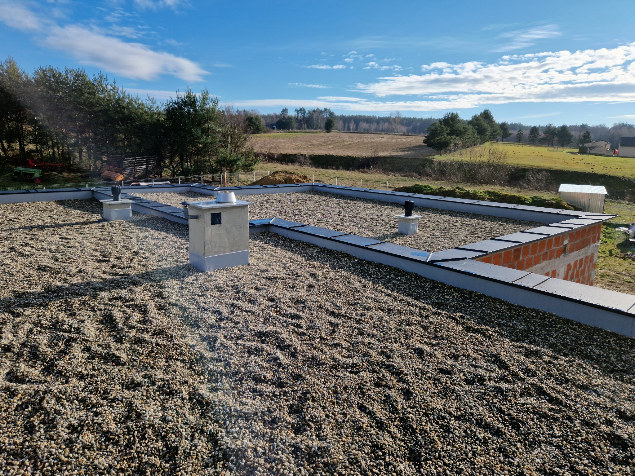 Płaski dach pokryty drobnym żwirem, obrzeża z blachy i fragment muru komina w surowym stanie na tle krajobrazu z drzewami i błękitnym niebem.