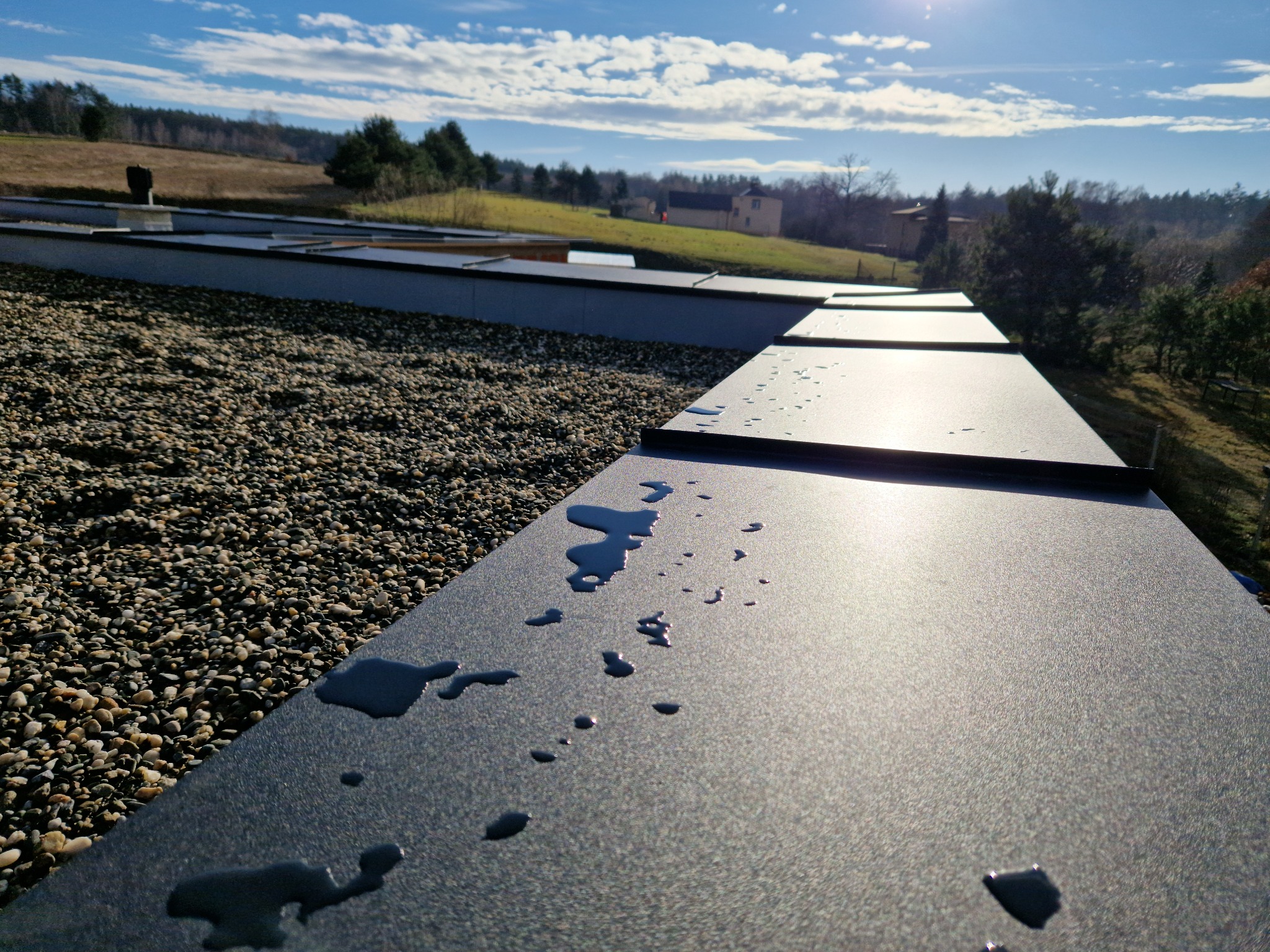 Płaski dach pokryty drobnym żwirem, z widocznymi panelami blachy trapezowej i kroplami wody, w tle zielone wzgórza i błękitne niebo.