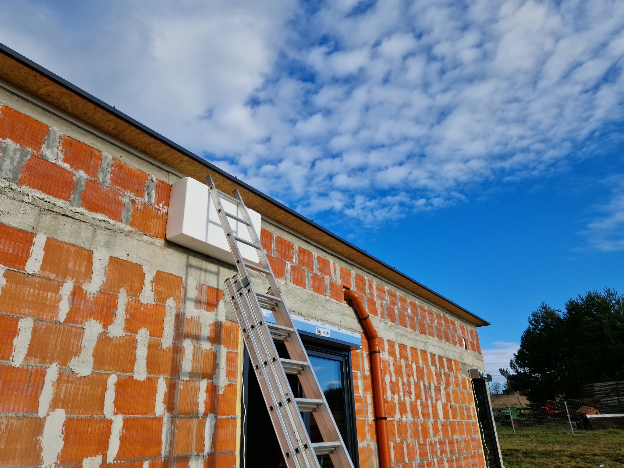 Budynek w trakcie budowy z niezakończonym dachem, widoczna drabina oparta o ścianę z pustaków, fragment ocieplenia i rura spustowa na tle błękitnego nieba.