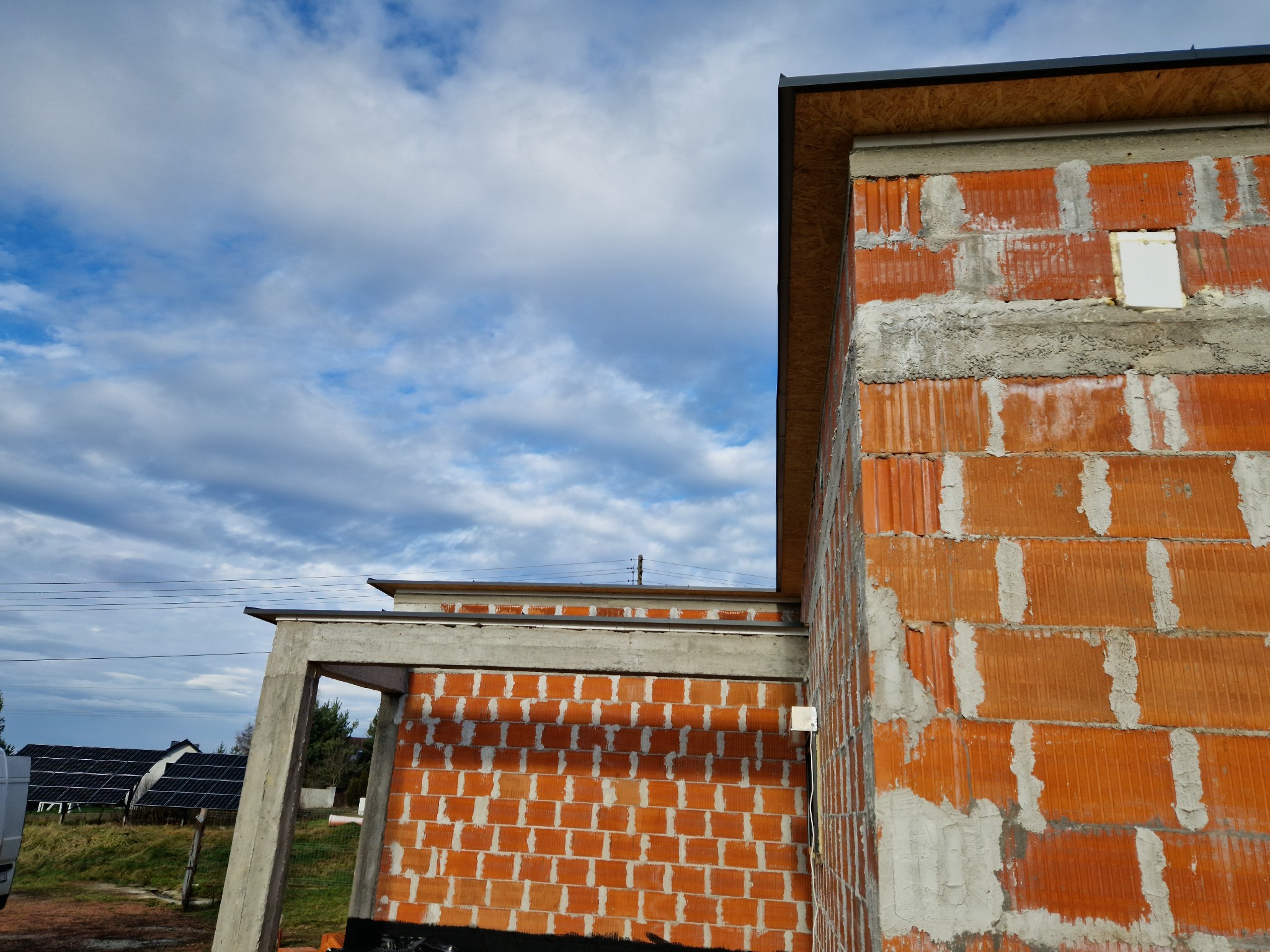 Surowy stan budynku z czerwonej cegły ceramicznej, widoczne elementy konstrukcji dachu z desek, w tle błękitne niebo z chmurami i panele słoneczne.