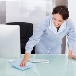 Uśmiechnięta kobieta w uniformie czyści szklany blat biurka niebieską ściereczką, obok monitor i klawiatura.