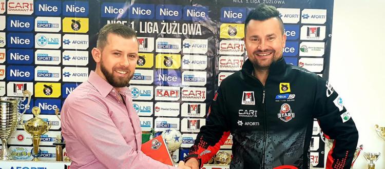 Dwóch mężczyzn w różnym wieku w sportowych strojach ściska sobie dłonie na tle ściany z logotypami sponsorów, przed nimi stoją puchary.