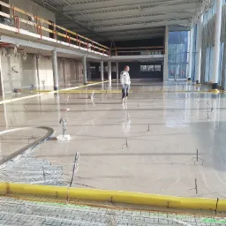 Świeżo wylana, mokra posadzka w dużym, jasnym pomieszczeniu z widocznym systemem ogrzewania podłogowego; pracownik w jasnym ubraniu stoi w oddali.