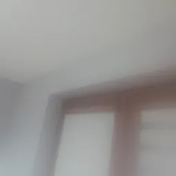 Fragment białej ściany i sufitu po nałożeniu gładzi, widoczne delikatne przejścia tonalne i struktura powierzchni. W tle widoczny fragment brązowej ramy okiennej.