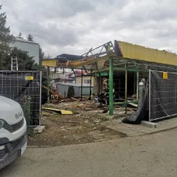 Demontaż konstrukcji z żółtym dachem, zabezpieczony siatką i tablicami ostrzegawczymi, z widocznym fragmentem białego samochodu dostawczego i butlą z gazem.