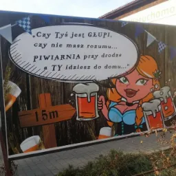 Tablica reklamowa piwiarni z ilustracją blondynki w stroju bawarskim trzymającej kufle piwa i napisem 'Czy Tyś jest GŁUPI, czy nie masz rozumu... PIWIARNIA przy drodze a TY idziesz do domu...'...