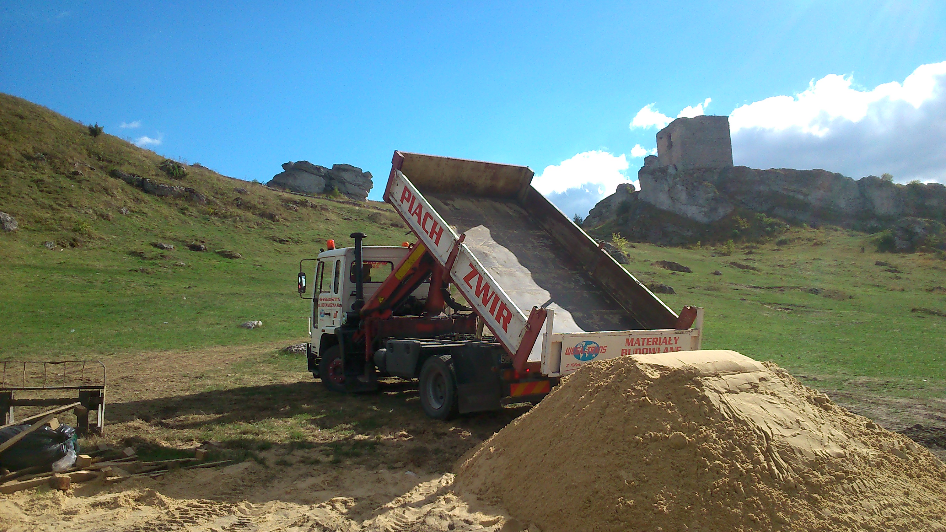 Wyładowany piasek z wywrotki na tle pagórkowatego krajobrazu z ruinami zamku w tle.