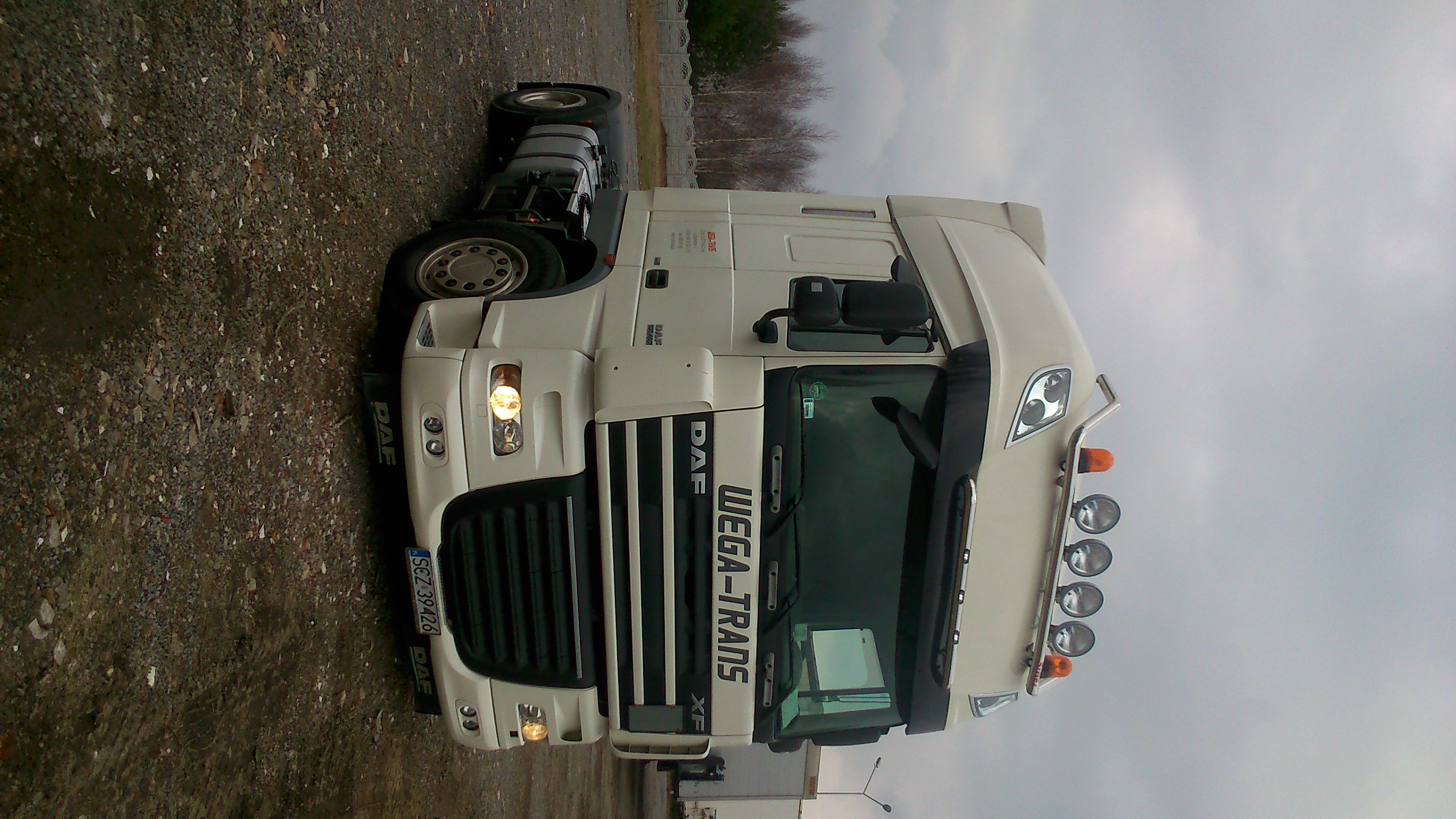 Biała ciężarówka DAF WEGA-TRANS XF zaparkowana na żwirowym terenie, widoczna z przodu, z włączonymi światłami mijania i dodatkowymi reflektorami na dachu, tablica rejestracyjna SCZ 39426.