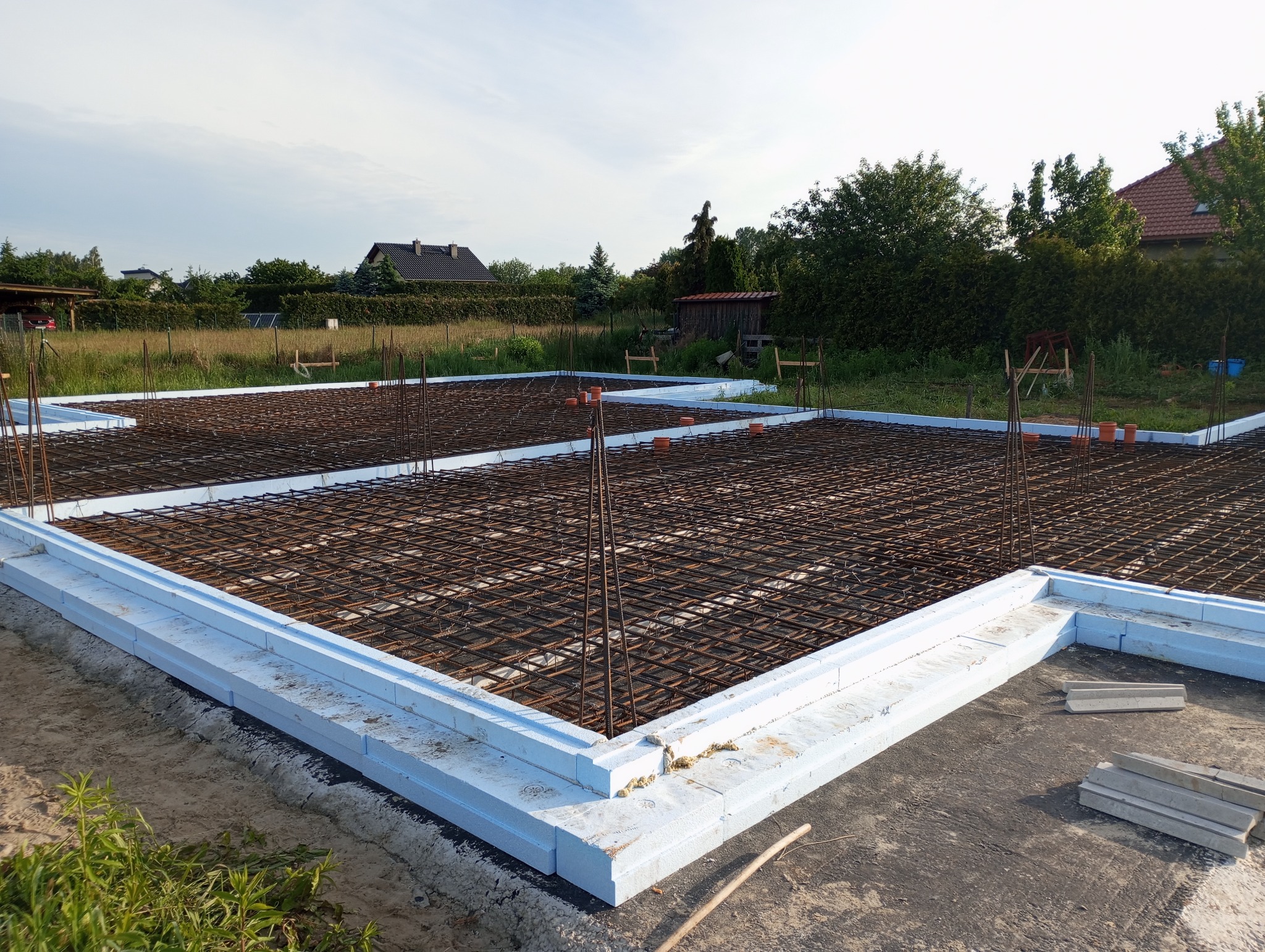 Zbrojenie fundamentów domu jednorodzinnego z użyciem białego szalunku styropianowego. Widok na siatkę zbrojeniową i wystające pręty. W tle zieleń i zabudowa.