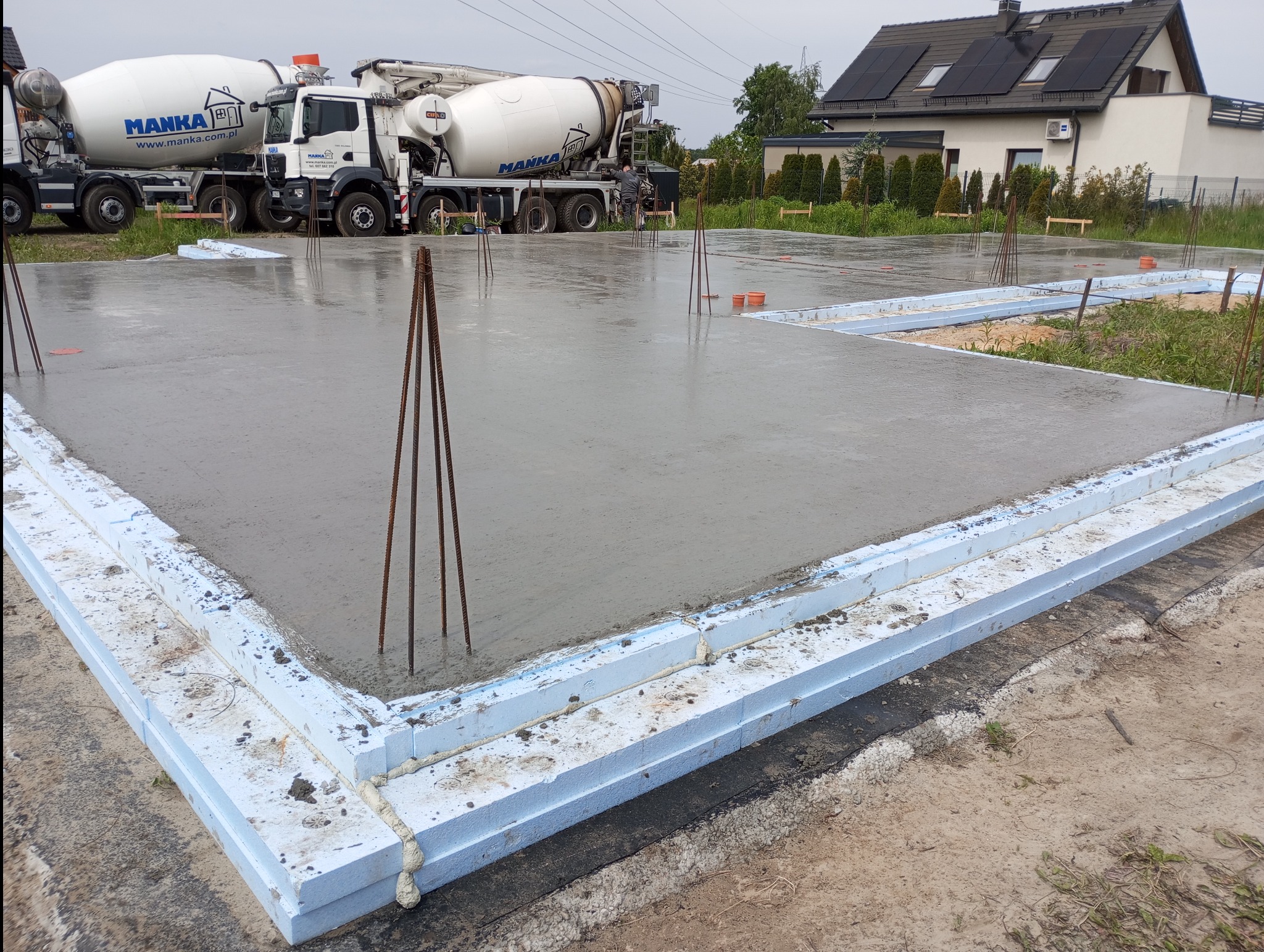 Wylana płyta fundamentowa z izolacją obwodową ze styropianu, widoczne wystające pręty zbrojeniowe i betonomieszarki w tle. Etap budowy domu jednorodzinnego.