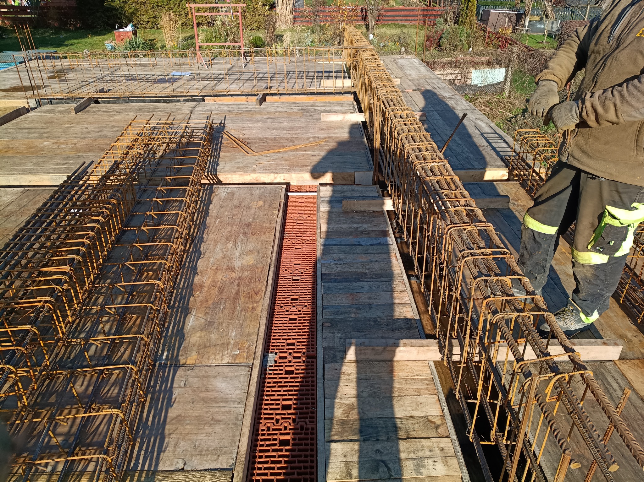 Zbrojenie stropu na deskowaniu, widoczne pręty zbrojeniowe ułożone w szkielet, ceglana kształtka wentylacyjna i fragment sylwetki pracownika w odzieży roboczej.