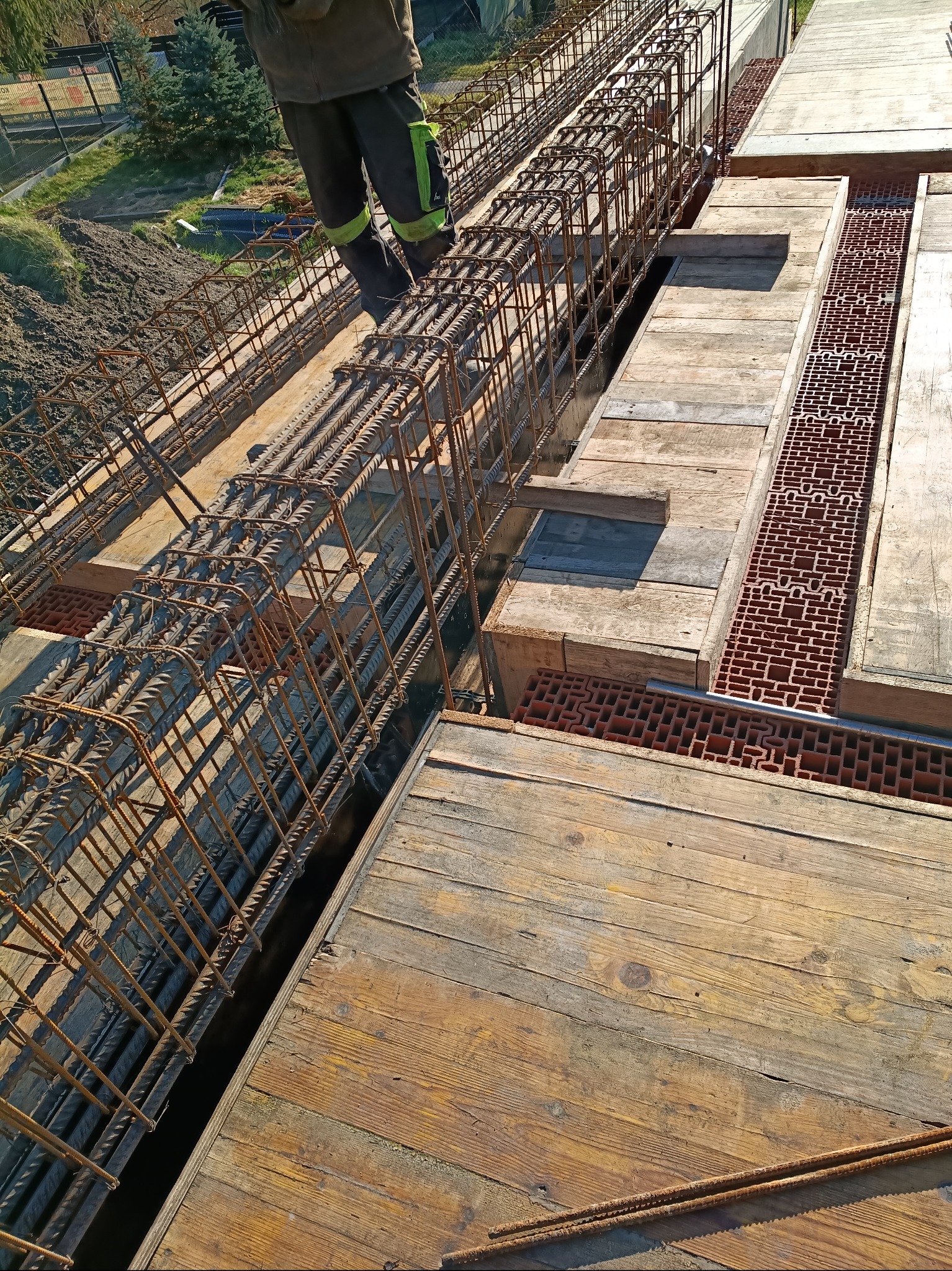 Zbrojenie fundamentów z widocznymi szalunkami i fragmentem pracownika w odzieży roboczej. Widok z góry na przygotowany teren pod wylewkę.