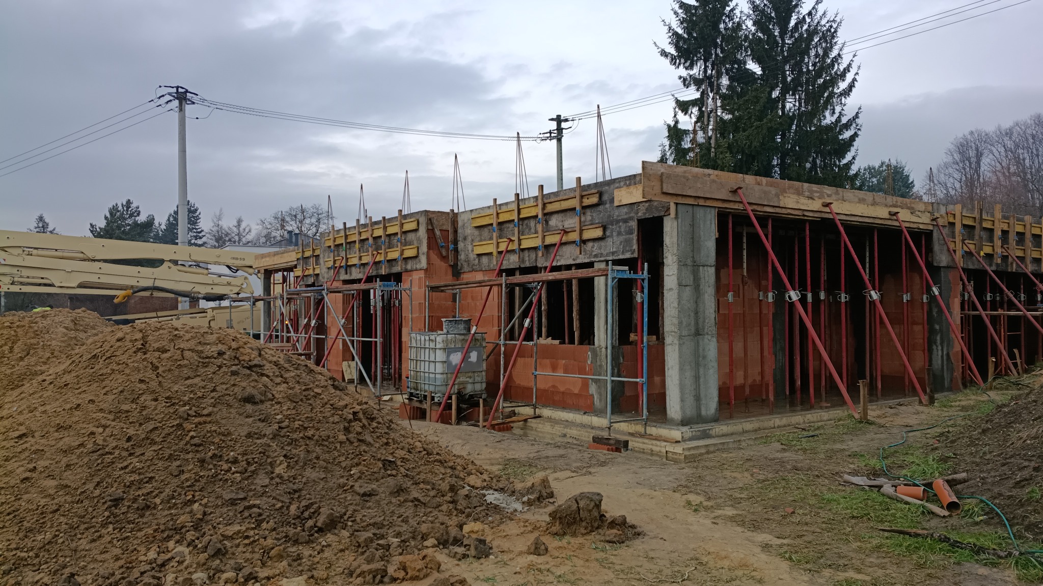 Wznoszenie ścian parterowego domu z czerwonej cegły, z widocznym szalunkiem i podporami. Na pierwszym planie hałda ziemi, w tle drzewa i słupy energetyczne.