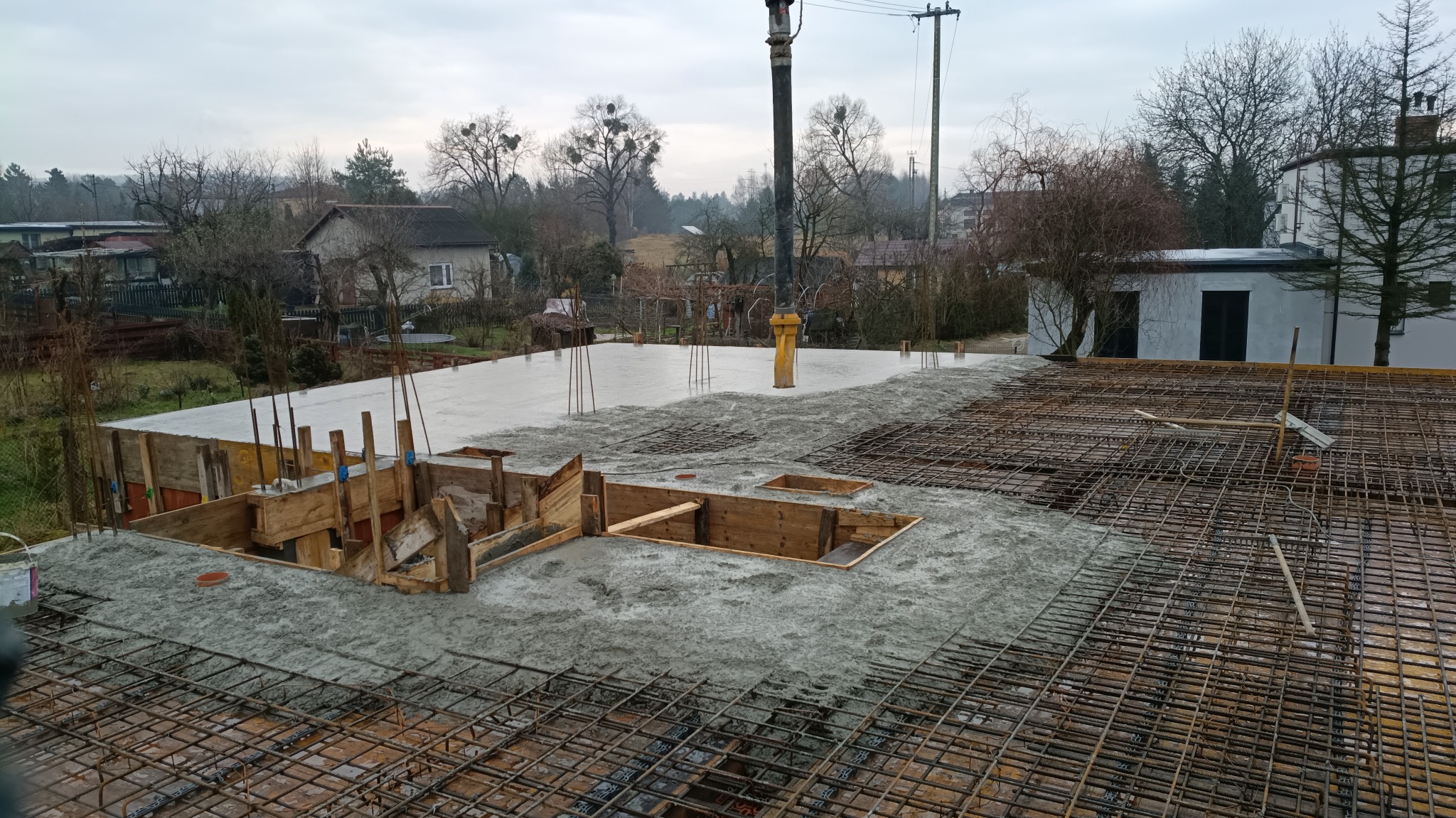Zalewanie betonem zbrojonej płyty fundamentowej domu jednorodzinnego. Widoczne szalunki i elementy konstrukcyjne. Prace w toku, pochmurny dzień.