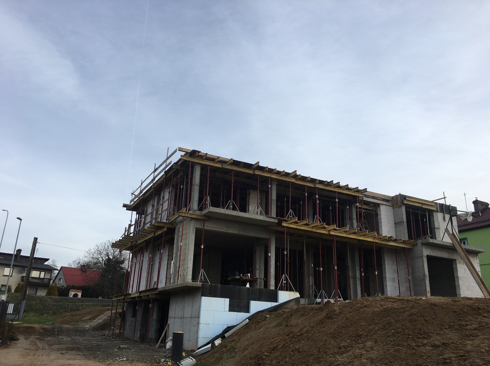 Surowy stan dwukondygnacyjnego domu z betonu komórkowego w trakcie budowy, z widocznym szalunkiem i ociepleniem fundamentów, na tle pochmurnego nieba.