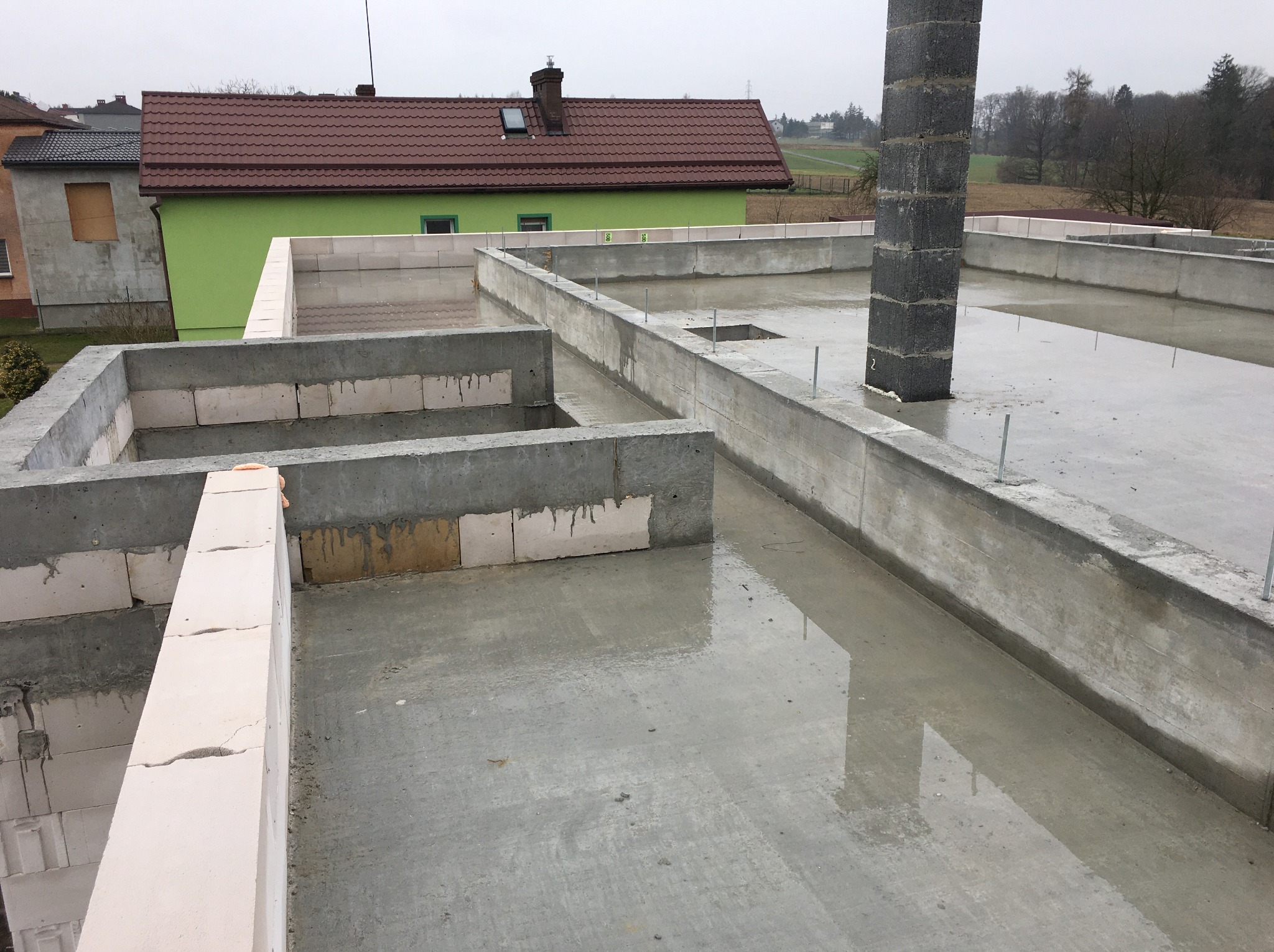 Wylany fundament domu z bloczków betonowych i słupem z pustaków szalunkowych, widok z góry na mokry beton, w tle zielony dom z brązowym dachem.