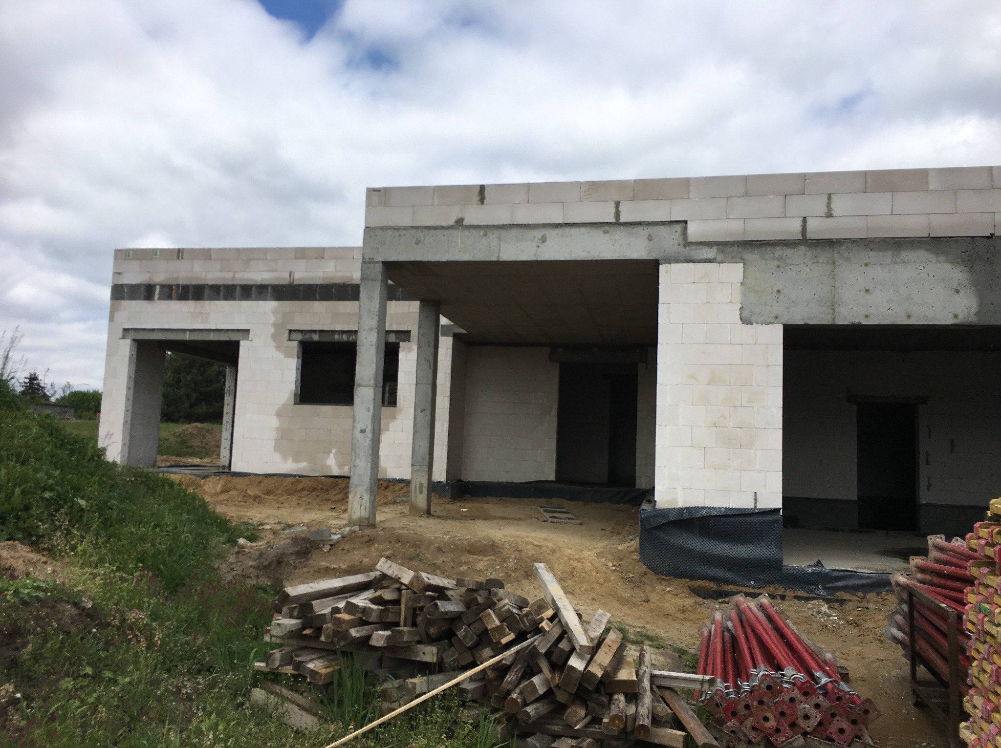Surowy stan domu z betonu komórkowego, widoczne szalunki i podpory. Budynek w trakcie wznoszenia, z otworami okiennymi i drzwiowymi. Na pierwszym planie drewniane i metalowe elementy.