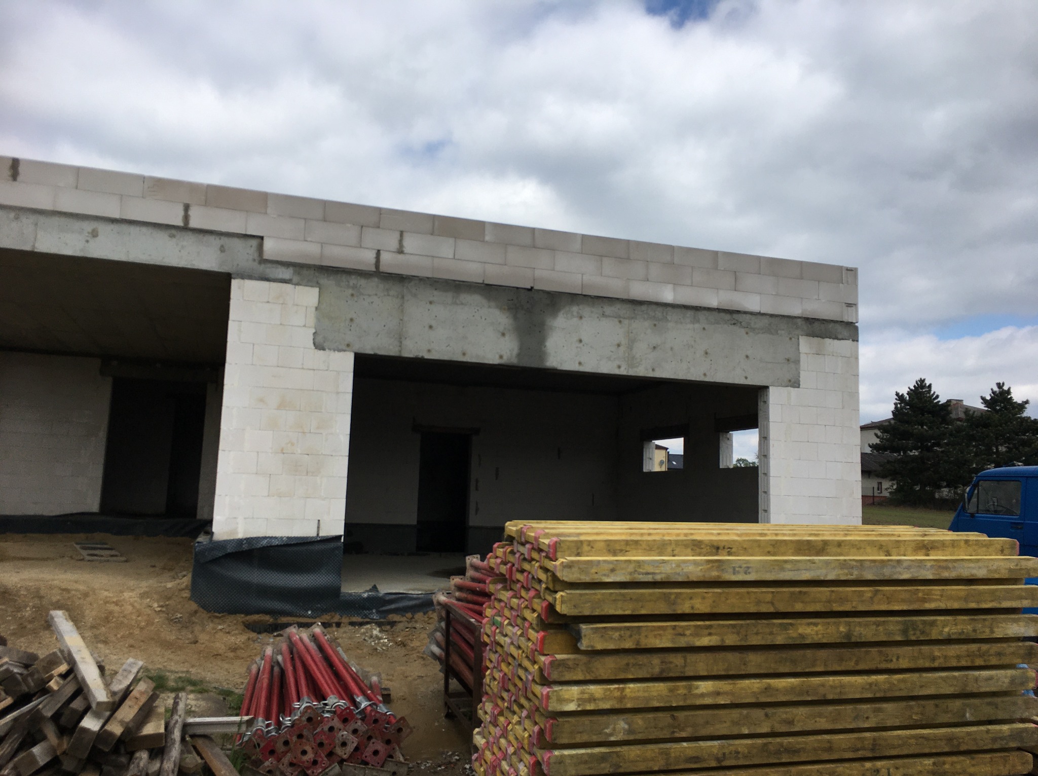 Surowy stan domu z betonu komórkowego, widoczne stemple i deski szalunkowe. Częściowo zadaszona konstrukcja w trakcie budowy, zarys okien i drzwi.