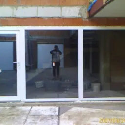 Białe okno PCV z drzwiami, widoczne od zewnątrz, z odbiciem osoby w środku, zamontowane w surowym murze z cegły.