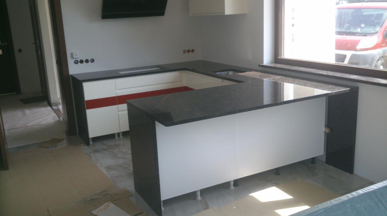 Nowoczesna kuchnia z blatem z ciemnego granitu i białymi szafkami, z widocznymi elementami wykończeniowymi i zabezpieczeniami podłogi podczas montażu.