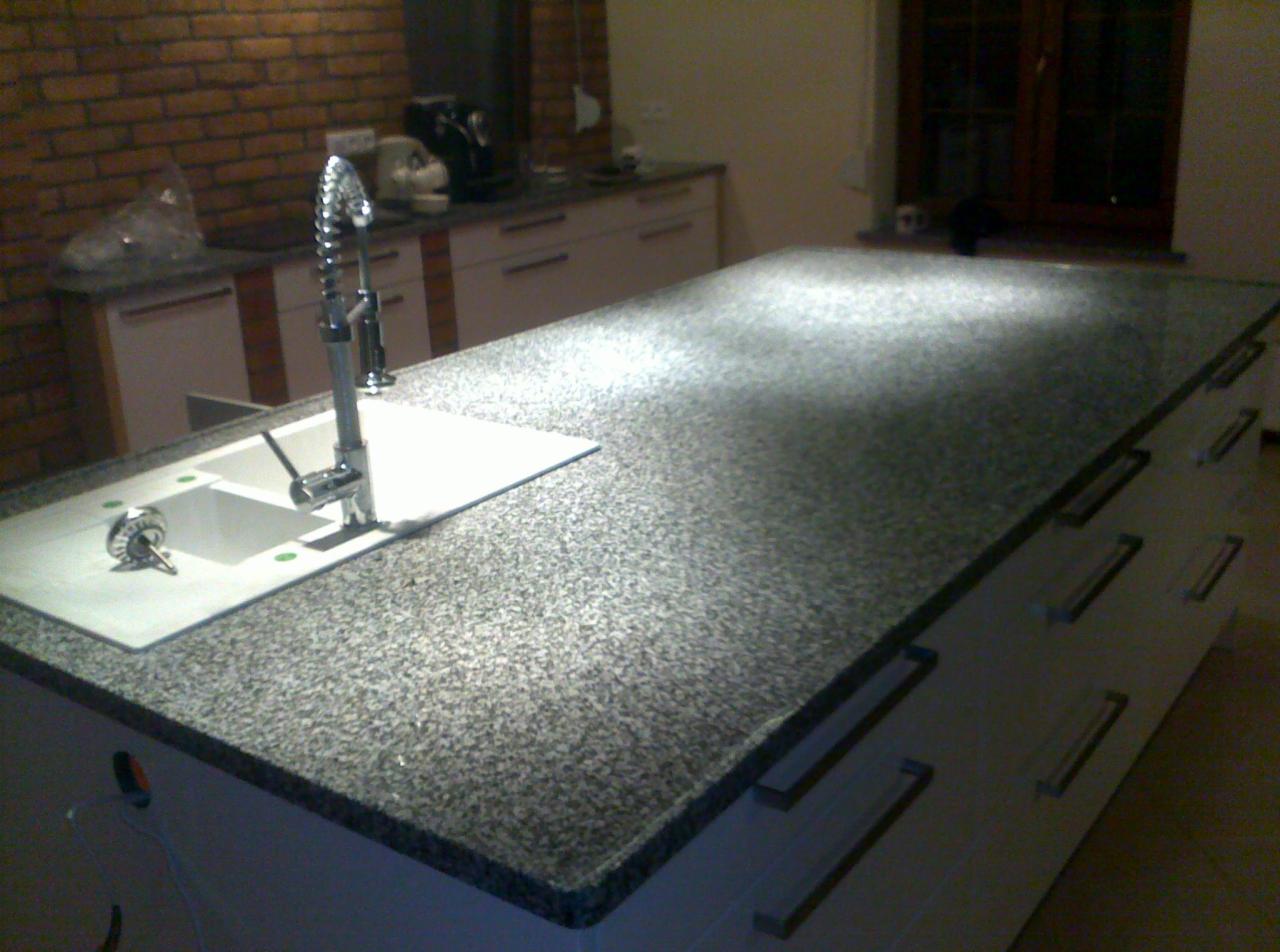 Szary, nakrapiany blat kuchenny z wbudowanym, białym zlewozmywakiem i nowoczesną baterią z wyciąganą wylewką, na tle ściany z cegły.