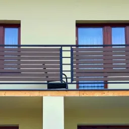 Nowoczesny balkon z brązowymi, poziomymi panelami i czarną metalową konstrukcją balustrady, widok z zewnątrz, z fragmentem drzewa w prawym górnym rogu.