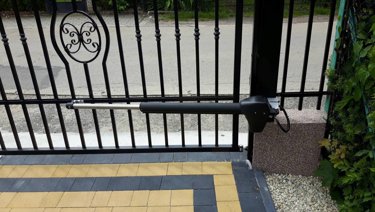 Automatyczna brama wjazdowa z czarnymi metalowymi prętami i dekoracyjnym elementem, widoczny siłownik napędu bramy.