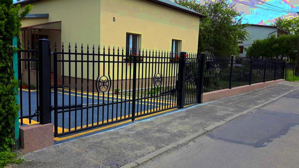 Czarna, metalowa brama wjazdowa z ozdobnymi elementami, widok z zewnątrz, na tle żółtego domu z numerem 5 i fragmentem ogrodzenia.