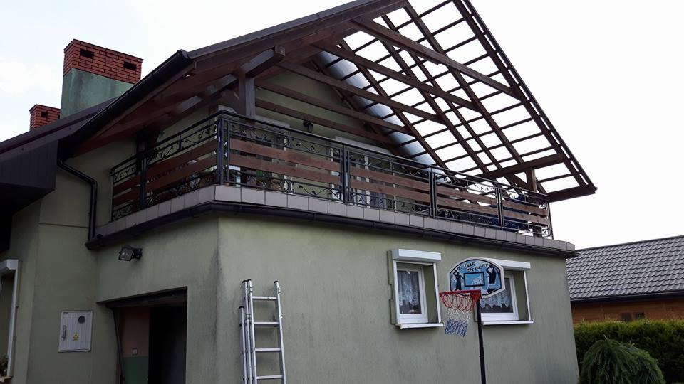 Zadaszony balkon z drewnianą konstrukcją i metalową balustradą z drewnianymi wypełnieniami, widok z zewnątrz budynku z drabiną opartą o ścianę i koszem do koszykówki zawieszonym nad oknem.