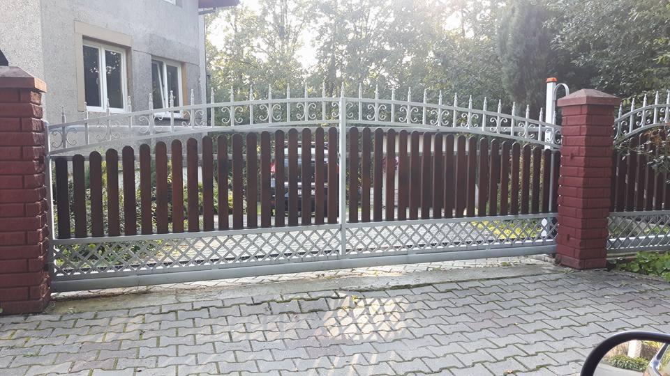 Dwuskrzydłowa brama wjazdowa z drewnianym wypełnieniem i metalową ramą, osadzona na murowanych słupkach z cegły klinkierowej, widok z zewnątrz na kostkę brukową i zieleń w tle.