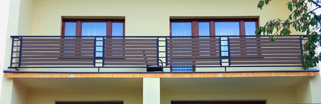 Nowoczesny balkon z brązowymi, poziomymi panelami i czarną metalową konstrukcją balustrady, widok z zewnątrz, z fragmentem drzewa w prawym górnym rogu.