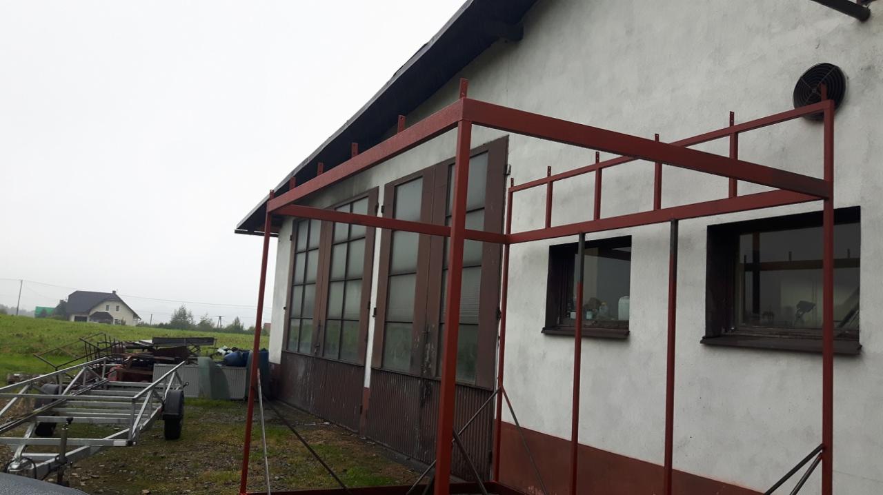 Metalowa konstrukcja w kolorze czerwonym, prawdopodobnie pod zabudowę balkonu lub tarasu, w trakcie montażu przy budynku z widocznymi oknami i wentylatorem na ścianie, na zewnątrz widoczny teren...