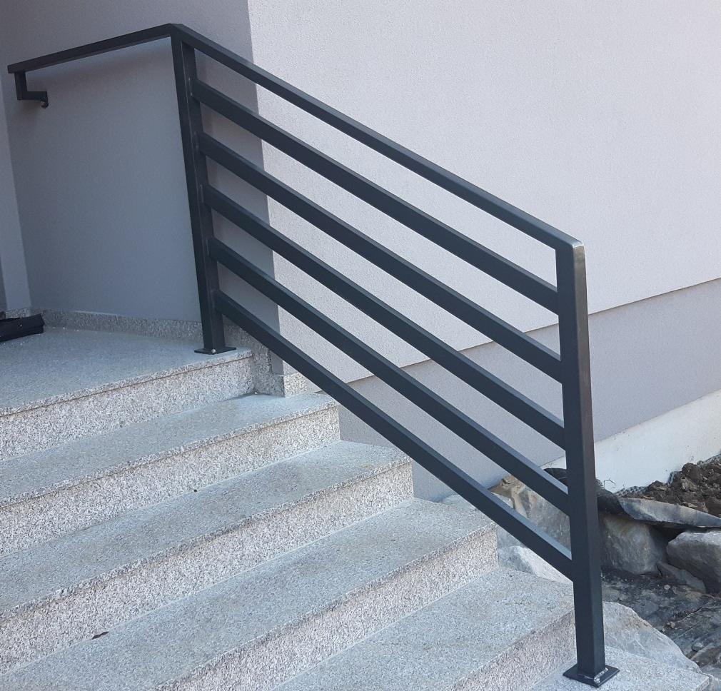 Zewnętrzne schody z szarego granitu z nowoczesną, minimalistyczną poręczą metalową z poziomymi szczeblami, mocowaną do ściany budynku.