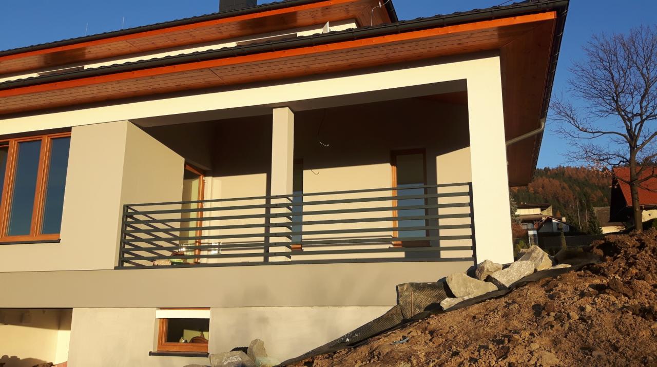 Nowoczesny dom z balkonem zabezpieczonym poziomą, metalową balustradą w kolorze grafitowym; widoczny nasyp ziemny z kamieniami u podstawy balkonu.