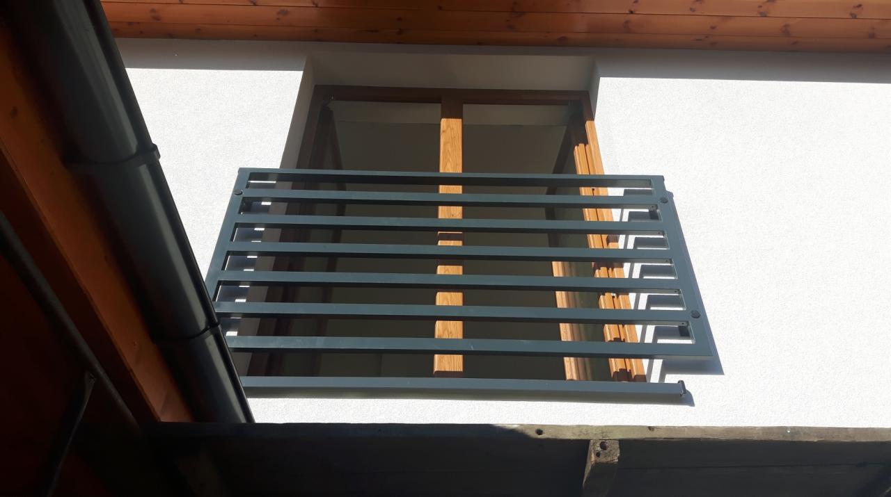 Szara metalowa balustrada balkonowa z poziomymi szczebelkami, zamontowana na białej elewacji budynku, widok z dołu, z fragmentem dachu w tle.