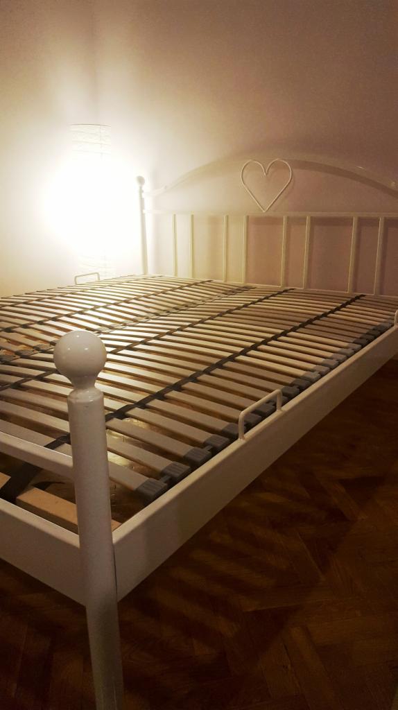 Białe metalowe łóżko z drewnianym stelażem i wezgłowiem z motywem serca, oświetlone lampą, na parkiecie.