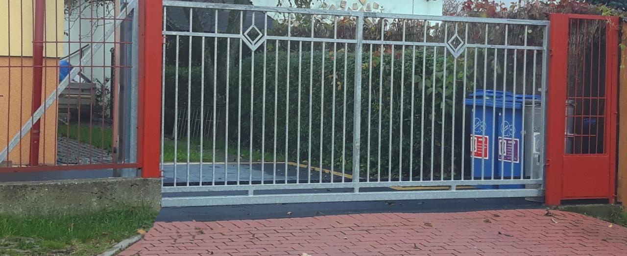 Przesuwna brama wjazdowa z metalowymi prętami i dekoracyjnymi rombami, pomalowana na szaro, z czerwonymi słupkami w Bielsku-Białej, widoczna na tle zieleni i niebieskich pojemników na śmieci.