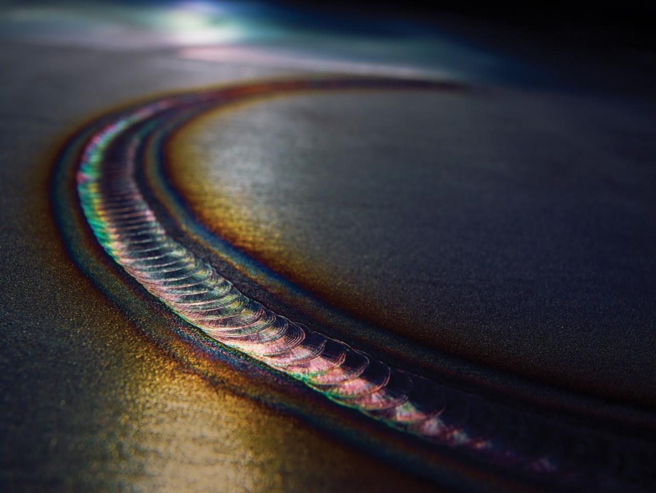 Zbliżenie na spaw metalu z widocznymi tęczowymi przebarwieniami termicznymi wzdłuż spoiny.