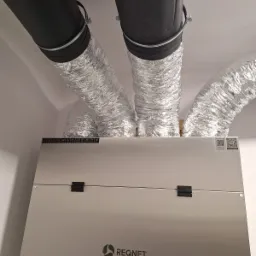 Centrala wentylacyjna z rekuperacją, widoczne czarne i srebrne rury wentylacyjne pod sufitem, zamontowana w pomieszczeniu.