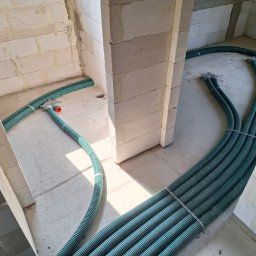 AIR+ Łomianki - Zielone, karbowane rury wentylacyjne ułożone na surowym betonie wewnątrz budynku z pustaków, widoczne elementy montażowe.