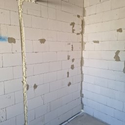 AIR+ Łomianki - Wewnętrzna ściana z betonu komórkowego w trakcie prac instalacyjnych, widoczne bruzdy wypełnione pianką montażową, rury instalacyjne prowadzone wzdłuż ścian i pod sufitem, surowy stan wykończenia.