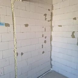 Wewnętrzna ściana z betonu komórkowego w trakcie prac instalacyjnych, widoczne bruzdy wypełnione pianką montażową, rury instalacyjne prowadzone wzdłuż ścian i pod sufitem, surowy stan wykończenia.