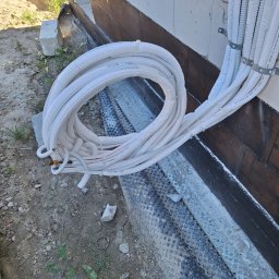 AIR+ Łomianki - Zwój białych rur klimatyzacyjnych leżący na ziemi obok fundamentu domu w trakcie instalacji.