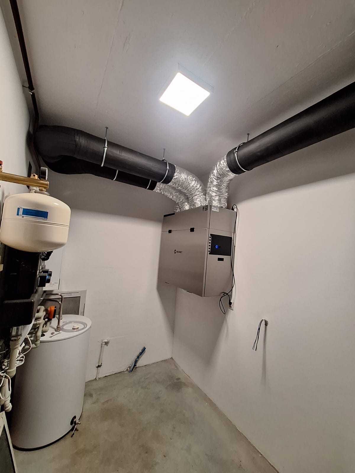 Zamontowana jednostka rekuperacji z czarnymi i srebrnymi przewodami wentylacyjnymi w jasnym pomieszczeniu technicznym, widoczne elementy instalacji grzewczej.