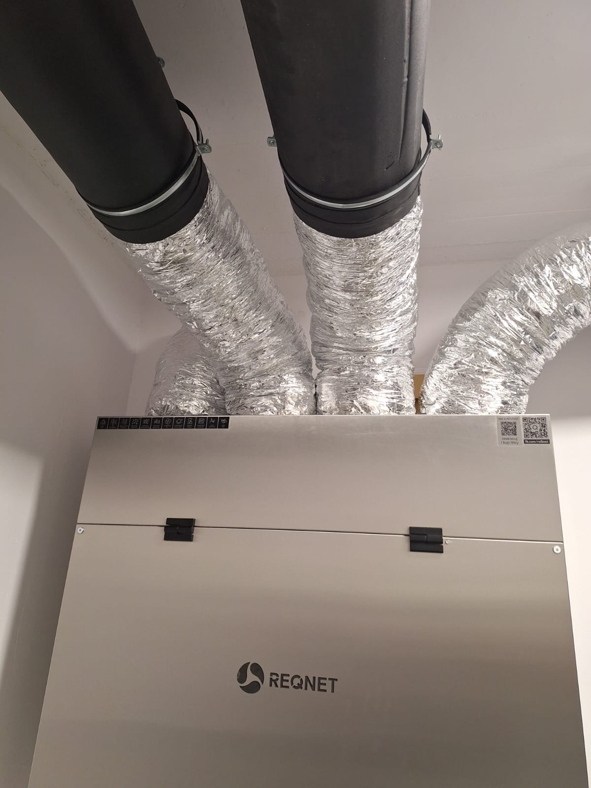 Centrala wentylacyjna z rekuperacją, widoczne czarne i srebrne rury wentylacyjne pod sufitem, zamontowana w pomieszczeniu.