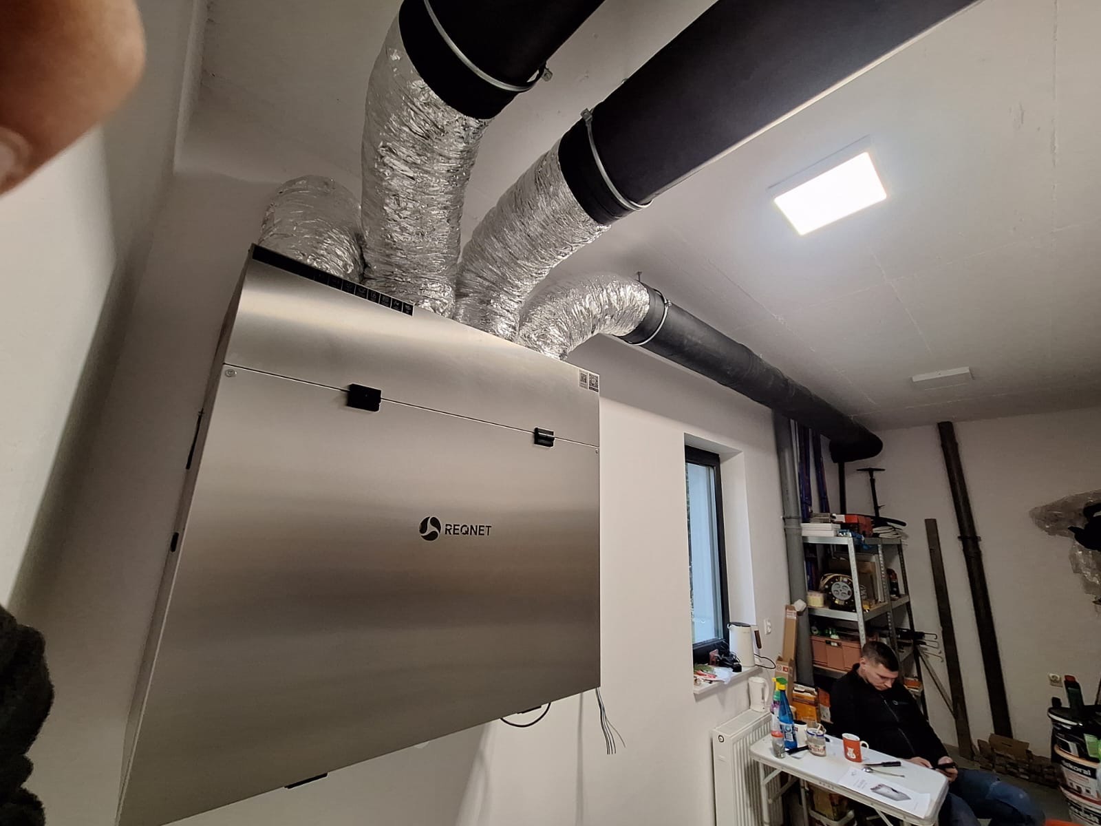 Srebrna centrala wentylacyjna z rekuperacją marki REQNET zamontowana w pomieszczeniu z widocznymi rurami wentylacyjnymi, oknem i siedzącym mężczyzną.