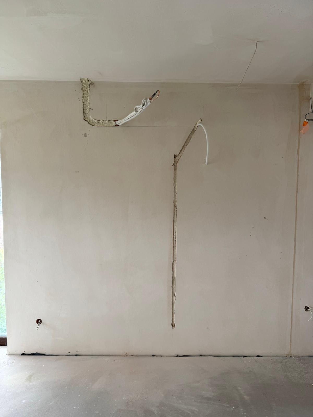 Ściana w trakcie prac instalacyjnych: widoczne bruzdy z przewodami, rura z izolacją piankową przy suficie, oraz otwory w dolnej części ściany.