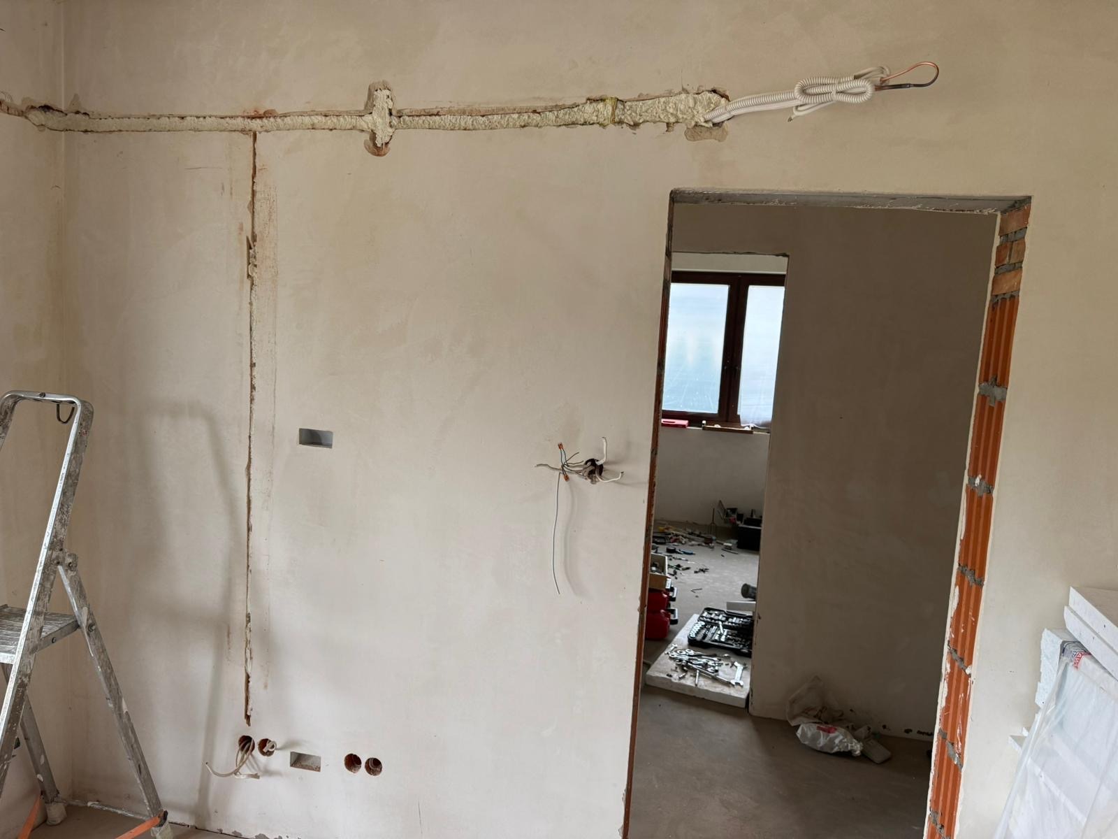 Wnętrze w trakcie remontu: przewody elektryczne zabezpieczone pianką montażową na ścianie, widoczne puszki elektryczne, drabina aluminiowa i narzędzia w tle.