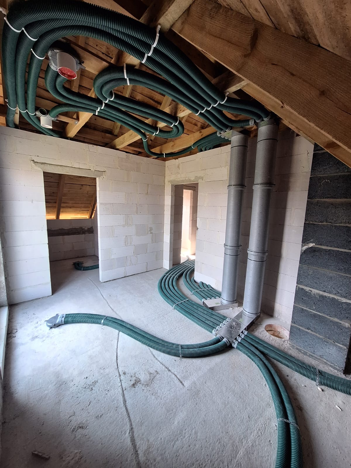 Zielone rury rekuperacji rozprowadzone pod drewnianym stropem na poddaszu w surowym stanie, widoczne szare rury pionowe i ściany z białego betonu komórkowego.