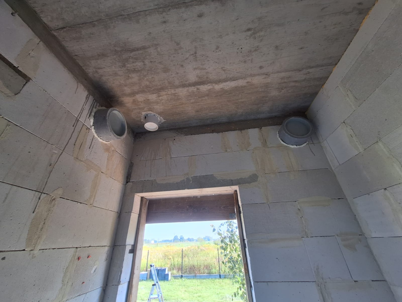 Surowy stan budynku z widocznymi otworami wentylacyjnymi w ścianach z betonu komórkowego i w suficie, widok przez otwarte drzwi na krajobraz za oknem.